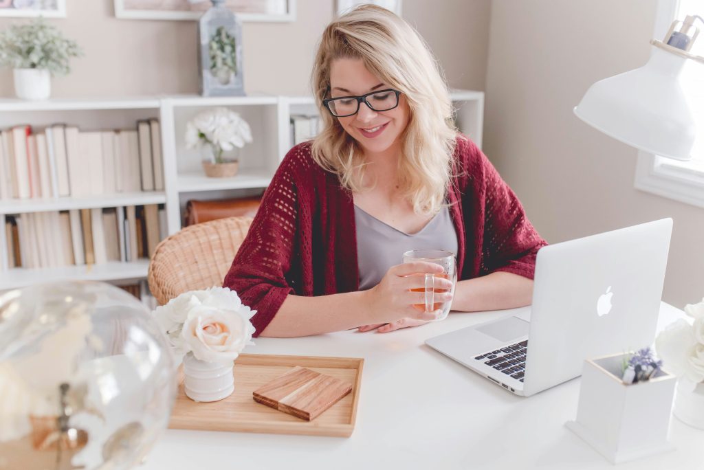 A woman sitting in front of a laptop, smiling, with a mug of tea in her hand.