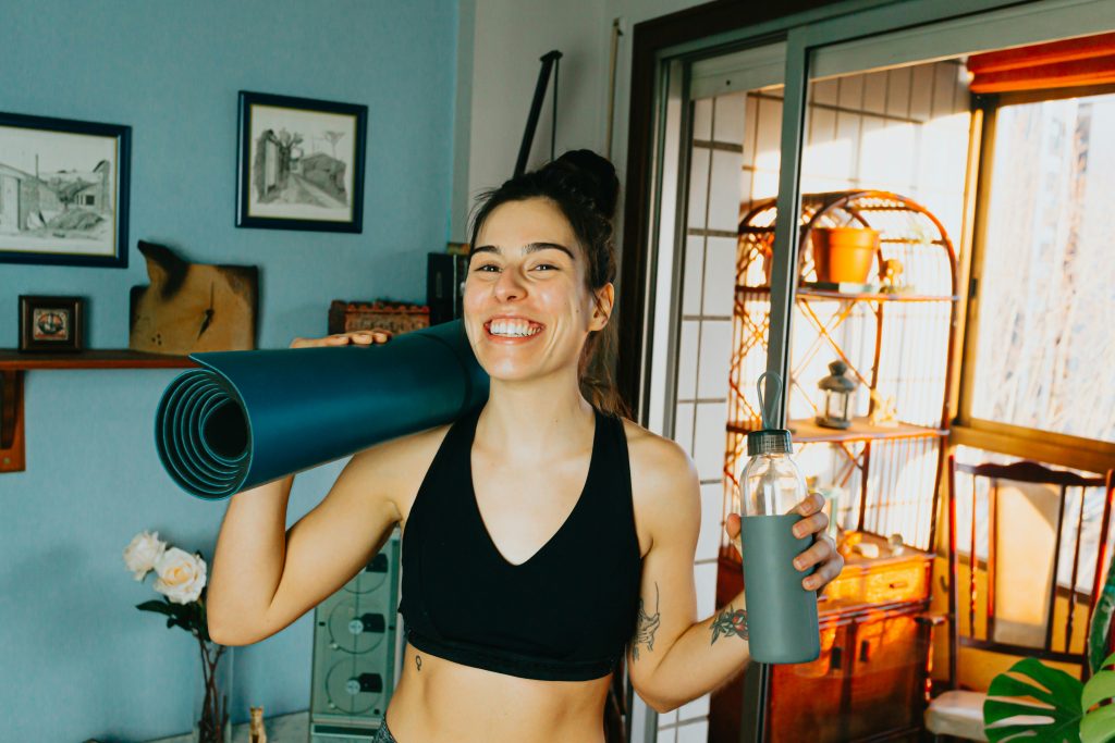 A woman in exercise clothing, holding a yoga mat and a water bottle, and smiling.