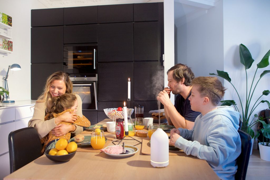A family sharing a meal around a table. There are candles lit on the table, and the mother is hugging her daughter.